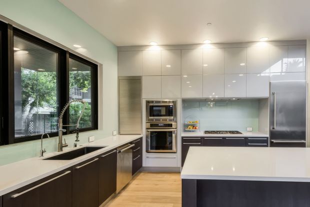 A bright contemporary kitchen with a large central island, dark lower cabinetry, glossy white full-height upper cabinets, pale glass splashback, integrated stainless-steel appliances, and light timber flooring. Large dark-framed windows bring in views of greenery, while recessed ceiling lights provide even illumination.