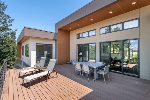 A contemporary two-storey-looking home exterior viewed from a spacious timber deck. The facade combines smooth light rendered walls, dark-framed floor-to-ceiling glazing, and warm timber soffit cladding. The deck includes outdoor dining furniture and loungers, with a railing and mature trees surrounding the property.