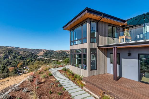 Contemporary multi-level residential home on a landscaped hillside, featuring vertical timber cladding, extensive floor-to-ceiling glazing, dark metal roof edges, and a timber deck with a concrete stepping-stone path. The view overlooks a dry, hilly landscape.