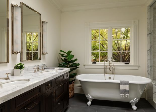 Elegant traditional bathroom with a dark wood double vanity, white marble countertop, polished metal fixtures, framed mirrors, a freestanding clawfoot bathtub, and a large divided-light window overlooking greenery. The room features detailed white trim and a dark tiled floor, creating a refined, bright, classic atmosphere.