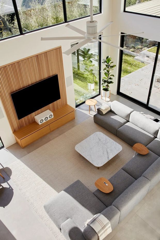 A bright, double-height living room viewed from above, with expansive black-framed floor-to-ceiling glazing, a large white ceiling fan, grey sectional sofa, pale rug, rounded marble-look coffee table, and warm timber media wall with vertical slatted panelling and built-in cabinetry. The room opens visually to a landscaped exterior through the glazing.