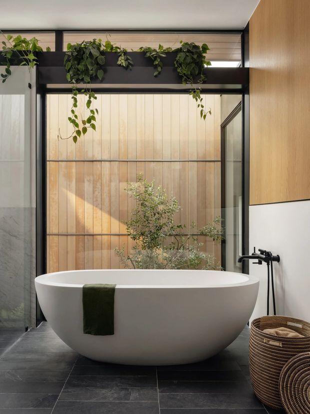 A serene contemporary bathroom centered on a large white freestanding oval bathtub. The room features dark stone-look tiled flooring, black-framed glazing, warm vertical timber wall panelling, a black wall-mounted bath filler, and abundant trailing greenery above the glazing. A planted courtyard or lightwell is visible beyond the window, creating a calm indoor-outdoor atmosphere.