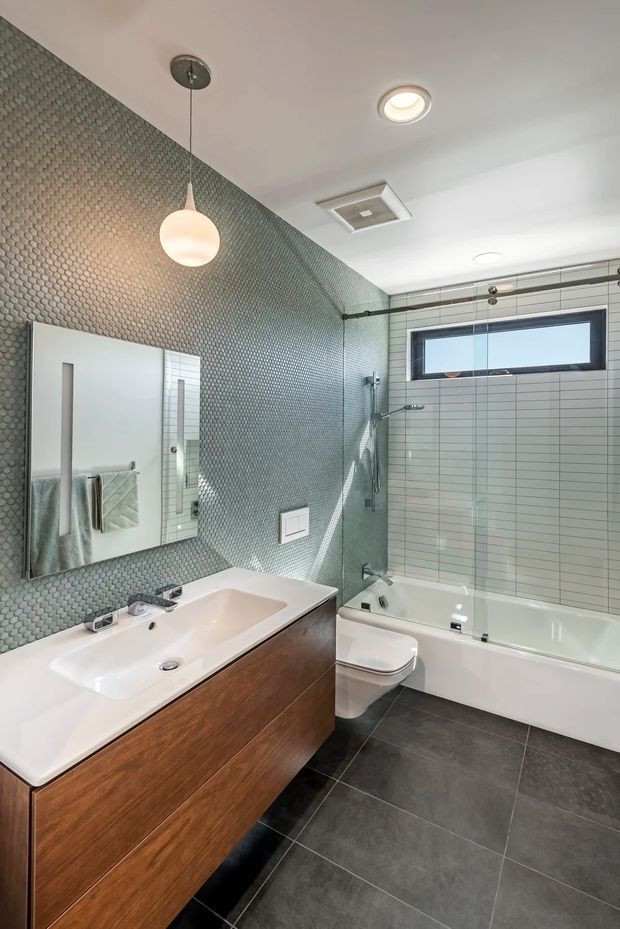 A contemporary bathroom featuring a wall-mounted timber vanity with a white integrated basin, mirrored cabinet, toilet, and a full-size bathtub with glass shower screen. The walls combine small green-grey hexagonal mosaic tiles with pale rectangular tiles, while the floor is finished in large dark grey tiles. A pendant light, recessed ceiling light, high horizontal window, and ceiling ventilation grille complete the space.