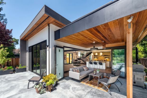 Modern single-storey home with a flat-roofed, open-plan indoor-outdoor entertaining area. The covered terrace features a warm timber-lined ceiling, exposed timber post, concrete paving, outdoor seating and an outdoor kitchen or bar connected to the contemporary interior. Dark metal cladding and expansive black-framed glazing contrast with white walls and natural timber finishes.