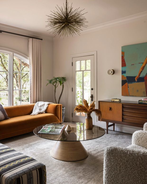 A refined living room with warm neutral walls, ornate ceiling moulding, tall arched windows, and a glazed exterior door. The room features a rust-orange sofa, textured cream armchair, patterned upholstered ottoman, round glass coffee table with a sculptural light-wood pedestal, mid-century timber sideboard, indoor plant, dried floral arrangement, and a dramatic spiky metallic pendant light. A colourful abstract artwork adds contrast.