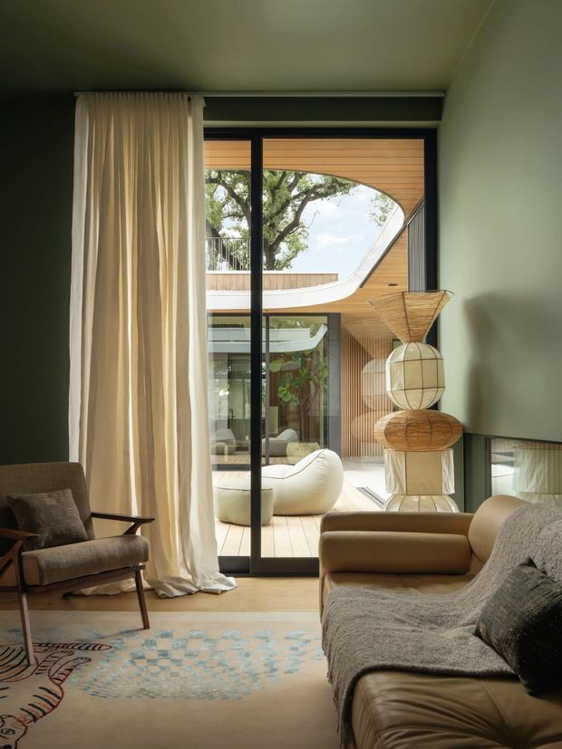 A warm, refined living room opening onto a landscaped courtyard through full-height black-framed glazing. The room features a low upholstered sofa, sculptural lounge chair, long cream curtains, muted green walls, a soft patterned rug, and an organic sculptural floor lamp. Outside, a timber-lined curved architectural form and pale decking are visible, with further seating in the courtyard.