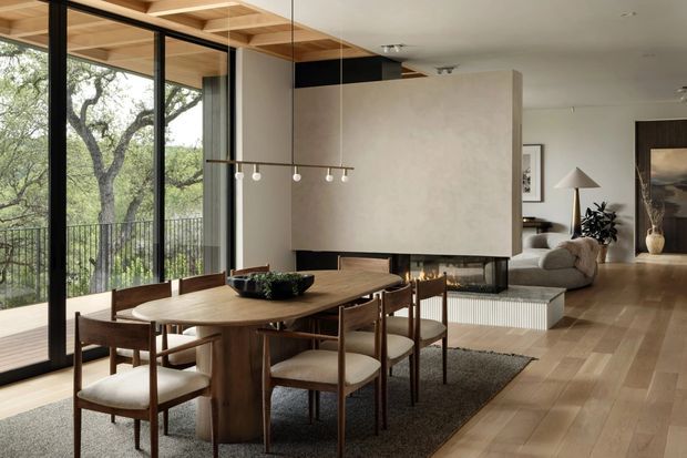 Open-plan dining and living area with a large timber dining table and mid-century-style chairs positioned beside full-height black-framed glazing. The room features warm timber flooring, a textured plaster fireplace wall with a linear firebox, a suspended multi-globe pendant, and a partially visible lounge with neutral furnishings. Mature trees and a balcony railing are visible beyond the glazing.