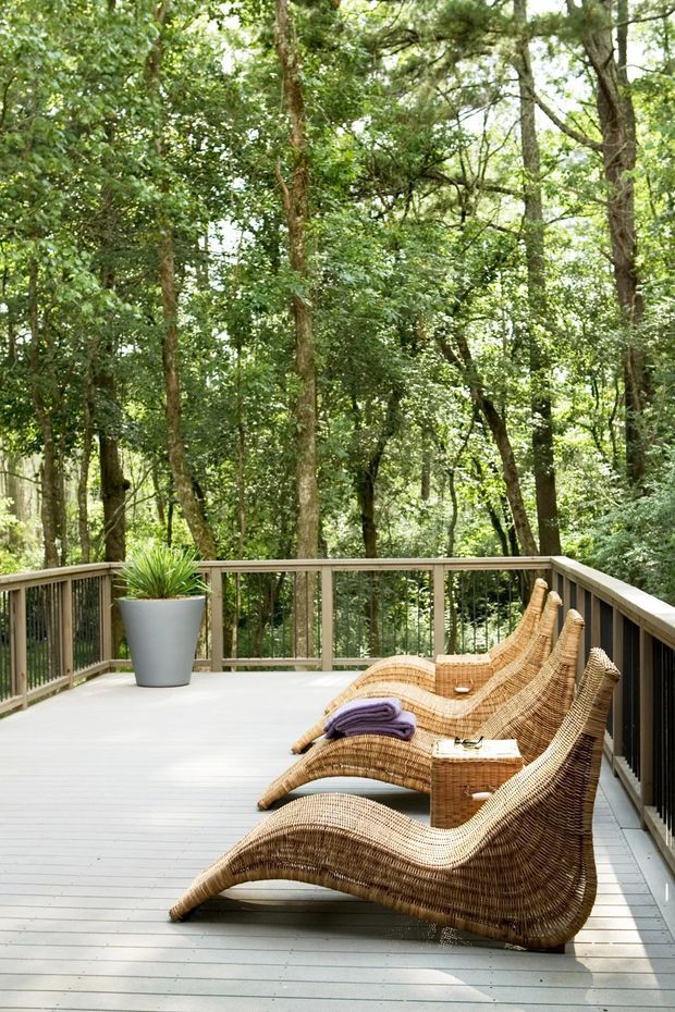 A spacious timber deck surrounded by dense woodland, furnished with a row of sculptural woven wicker chaise lounges. The deck has pale grey floorboards, timber handrails with dark vertical balusters, and a large grey planter with a green plant. The setting is bright, leafy, and tranquil, with the outdoor seating oriented toward the trees.