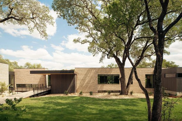 Single-storey contemporary residential building viewed from a landscaped lawn, with a long low-profile tan masonry facade, dark recessed entry and garage-like volumes, expansive horizontal glazing, and a gently sloping accessible walkway. Mature trees frame the building and cast dappled shadows across the lawn and facade.