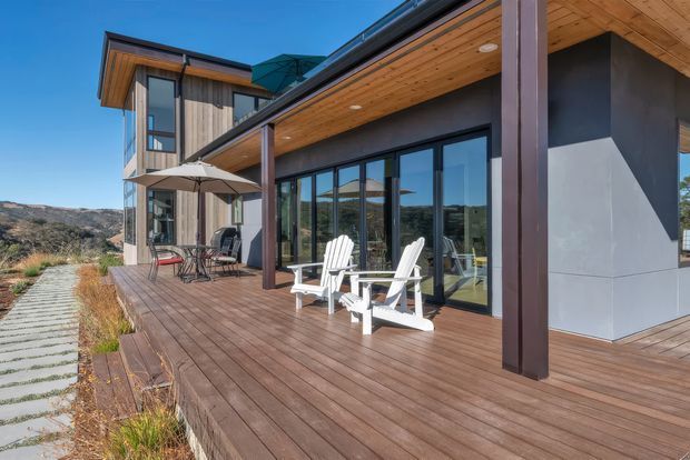 Modern multi-level home with a broad timber deck and covered outdoor living area. Dark vertical structural posts frame extensive floor-to-ceiling glazing, while timber soffits and timber cladding add warmth. White outdoor lounge chairs and a dining setting overlook a dry, rugged landscape.