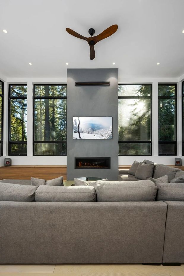 Contemporary living room with a large grey sectional sofa facing a central concrete-look fireplace wall with a built-in linear firebox and mounted television. Floor-to-ceiling black-framed windows surround the room, looking onto a wooded landscape. The space has a white ceiling with recessed downlights, warm timber flooring, and a sculptural three-blade timber ceiling fan.