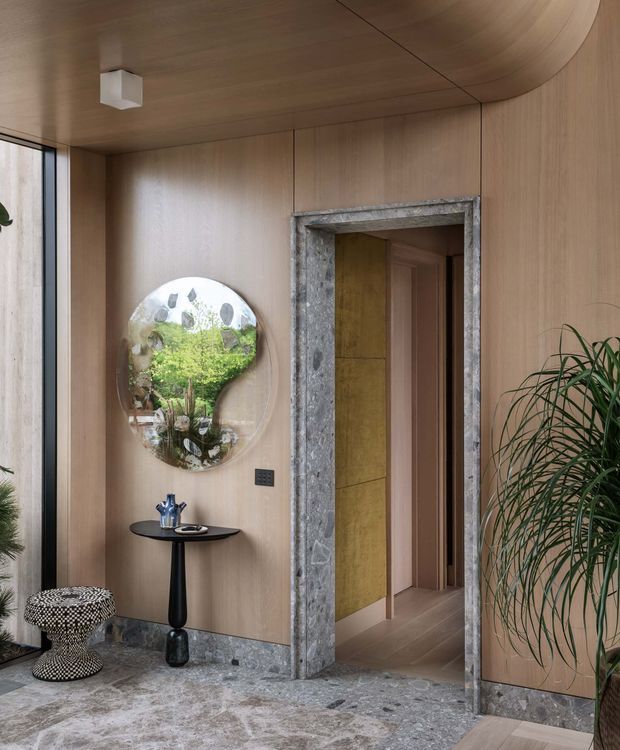 A refined interior entryway with warm timber wall and ceiling panelling, a curved ceiling detail, and a dramatic grey veined stone floor and door surround. A small black pedestal console with a decorative vessel sits beneath an irregular rounded mirror, while tall indoor plants and a patterned stool add texture. The open doorway leads to an adjoining corridor or room with additional timber finishes.