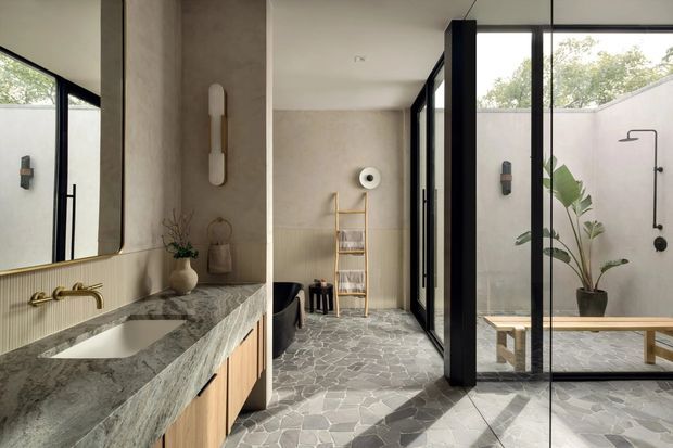 A refined contemporary bathroom with an open, courtyard-oriented layout. The foreground features a long marble-look vanity with an integrated basin, timber cabinetry, brass tapware, and a large mirror. A freestanding bathtub sits beyond a textured wall, while black-framed full-height glazing opens visually to a private courtyard with an outdoor shower and bench. Irregular grey stone flooring, warm neutral plaster, and indoor planting create a calm, spa-like atmosphere.