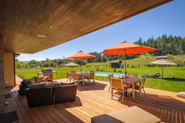 A spacious covered outdoor entertaining deck with timber flooring and a timber-lined ceiling, furnished with wicker lounge seating and outdoor dining tables beneath bright orange and neutral umbrellas. The deck opens onto a manicured lawn and landscaped rural garden with vineyards and trees beyond.