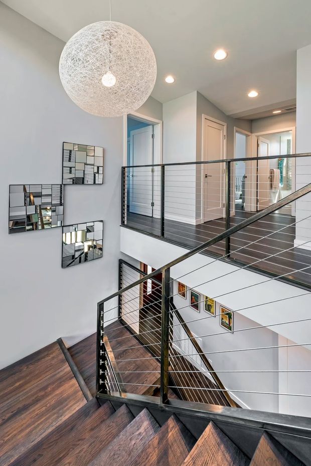 A bright multi-level residential stairway and landing viewed from the staircase. Dark stained timber stairs and flooring contrast with white walls and ceilings. Black metal railings with horizontal cable infill surround the stairwell, which features a tall open void and a large spherical woven pendant light. Several white paneled doors, recessed ceiling lights, framed artwork, and mirrored wall decor are visible.