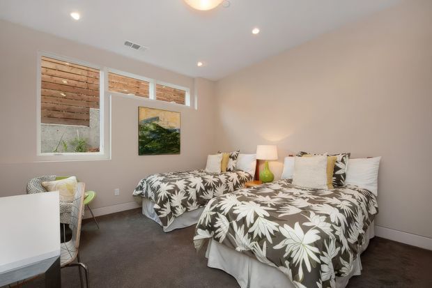 A bright, compact bedroom with two twin beds dressed in matching botanical-patterned grey and white bedding. The room has pale neutral walls, dark carpet, white trim, recessed ceiling lights, bedside lamps, and high windows with a timber privacy screen visible outside. A small desk and chair sit near the window, with framed artwork between the beds.