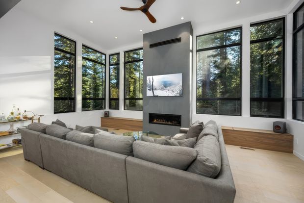 Bright contemporary living room with a large grey sectional sofa arranged around a slim built-in fireplace and wall-mounted television. Floor-to-ceiling black-framed windows wrap around the room, overlooking dense trees. The space has pale flooring, white walls, recessed lighting, a timber built-in console, and a ceiling fan.