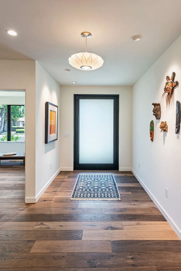 A bright residential entry hall with white walls, dark-framed frosted-glass front door, wide-plank timber flooring, and a patterned runner rug. Decorative carved masks and wall art line the right wall, while a sculptural pendant light hangs centrally from the ceiling. An adjoining living area is partially visible to the left.