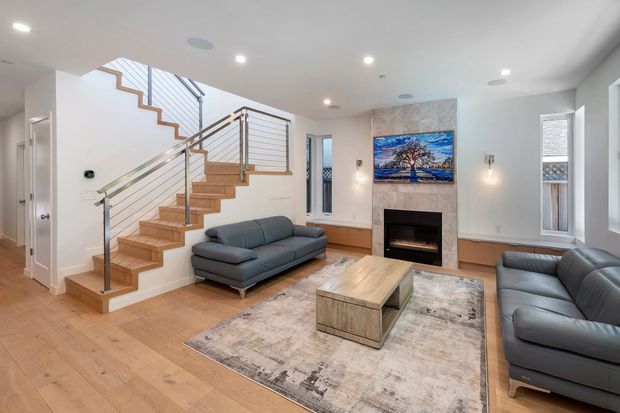 Bright contemporary living room with an open staircase, pale timber flooring, grey sofas, a large area rug, a timber coffee table, and a tiled fireplace feature wall with a mounted artwork. The staircase has timber treads, slim metal balusters, and a timber handrail. Windows and recessed ceiling lights provide ample natural and artificial light.
