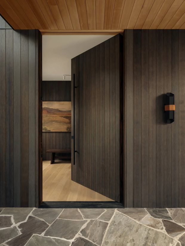 A dark, richly detailed interior threshold with a tall pivot door opening into a timber-lined room. Vertical dark wood wall panelling surrounds the doorway, while the ceiling is clad in warm timber boards. The foreground floor is irregular natural stone paving, contrasting with the pale timber floor beyond. A small wall light with a warm wood-toned accent is mounted on the right wall, and artwork and a bench are visible through the doorway.