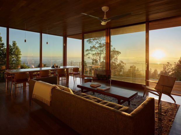 A warm, open-plan living and dining room with expansive floor-to-ceiling glazing overlooking a distant landscape at sunset. The room features dark timber flooring and a timber-lined ceiling, slender timber-framed windows, a long dining table, mid-century-style lounge furniture, a low coffee table, and a ceiling fan. The glazing and open layout create a strong indoor–outdoor connection and panoramic outlook.