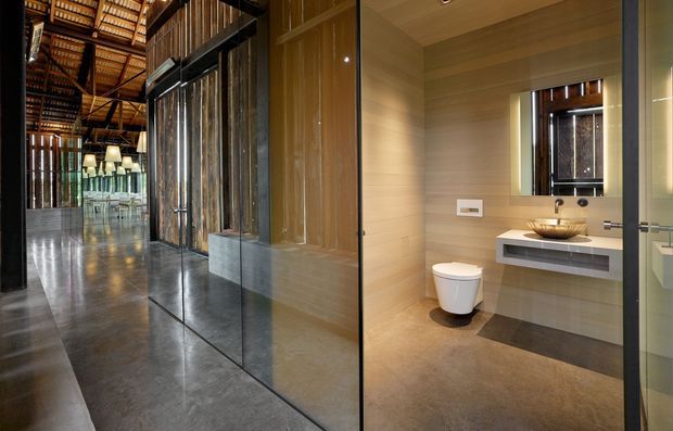 A contemporary bathroom viewed from an open glass doorway, featuring a wall-mounted toilet, floating vanity with a vessel basin, large mirror, and warm wood-toned wall and ceiling finishes. The bathroom opens directly onto a spacious polished concrete-floored interior with dark-framed glass partitions, exposed timber roof structure, and pendant lighting.