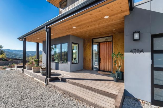 Contemporary residential house exterior viewed from the front porch, featuring a broad timber-lined covered entry, dark structural posts, grey rendered walls, a vertical timber front door, timber decking and steps, and large windows. Gravel landscaping and distant hills are visible beyond the porch.