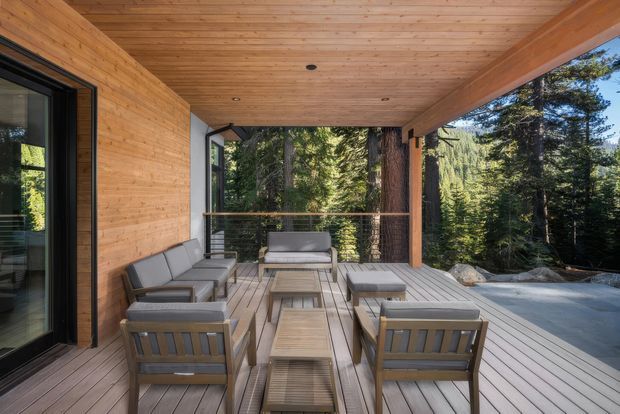 Covered outdoor terrace with timber decking, timber-clad walls and ceiling, and a substantial timber beam framing views into a dense forest. The terrace is furnished with a grey outdoor sofa, armchairs, and a low timber coffee table, with glazed sliding doors connecting to the interior and a glass balustrade along the edge. The design has a warm, contemporary cabin-like character and appears integrated with a wooded landscape.