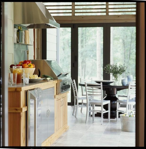 A bright, rustic kitchen and dining area in a timber-framed home. The kitchen features natural timber cabinetry, a pale stone or tiled floor, stainless-steel appliances, open shelving, and a large metal rangehood. Beyond the kitchen is a round dark dining table with white chairs, set beside expansive dark-framed glazed doors and windows overlooking a wooded landscape. Timber ceiling beams and clerestory glazing add warmth and natural light.
