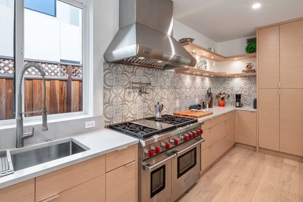 Bright, contemporary kitchen with light timber cabinetry, a large stainless-steel range cooker and hood, white benchtops, and an ornate patterned tile splashback. Open shelving displays ceramics and accessories, while a farmhouse-style sink sits beneath a large window overlooking a fenced exterior.