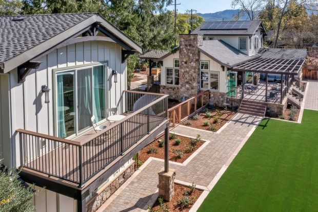 Elevated exterior view of a multi-level residential property with several connected pitched-roof buildings, grey weatherboard cladding, stone veneer accents, solar panels, covered outdoor areas, decks with dark metal balustrades, paved walkways, landscaped garden beds, and a bright green lawn. Mountainous terrain is visible in the background.