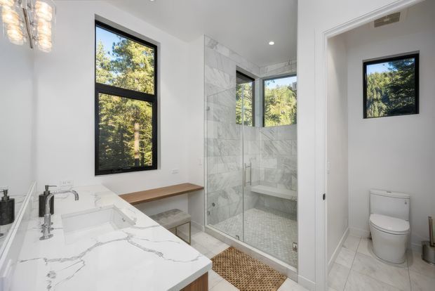 Bright contemporary bathroom with a white marble-look double vanity and polished chrome fixtures in the foreground. A glass-enclosed walk-in shower with large white veined wall tiles is centered, while a separate toilet room is visible to the right. Black-framed windows overlook leafy trees, and a woven bath mat adds warmth.