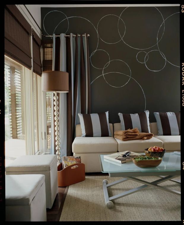 A refined living room with a large cream sectional sofa, striped brown-and-white cushions, a glass-topped coffee table with a metal geometric base, and a neutral textured rug. The room features tall windows and glazed doors with timber blinds and layered curtains, a sculptural floor lamp, and a dark feature wall decorated with oversized overlapping circular line artwork. The palette is warm and neutral with strong brown, cream, and charcoal contrasts.