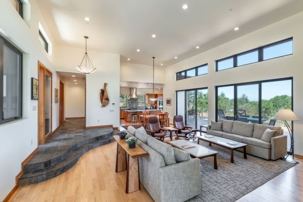 Bright open-plan living room with a double-height ceiling, large clerestory and full-height windows, a stepped stone entry platform, timber flooring, contemporary grey seating, a dining area, and a kitchen visible beyond. The room has abundant natural light and a refined neutral palette with warm timber accents.