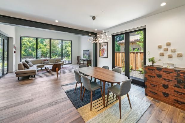 Open-plan interior combining a dining area and living room. The space has pale wide-plank timber flooring, white walls and ceiling, extensive black-framed glazing and sliding doors, a large rectangular timber dining table with upholstered chairs, and a relaxed living area with a sofa and lounge chairs. A patterned rug anchors the dining zone, while a timber chest of drawers and wall artwork add warmth. The room is bright, contemporary, and connected to a small leafy outdoor courtyard through the glazing.