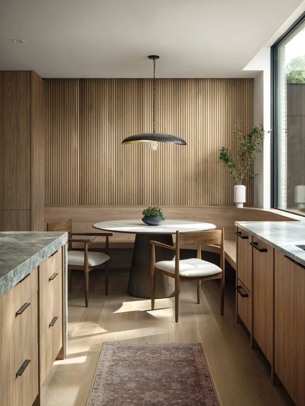 Warm, refined kitchen-dining interior with extensive mid-tone timber cabinetry and a vertical timber-slat feature wall. A round dining table with upholstered wooden chairs sits in a built-in banquette beside large floor-to-ceiling glazing. Stone-topped kitchen islands frame the foreground, while a sculptural pendant light and soft natural daylight create an elegant, inviting atmosphere.