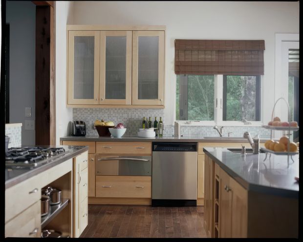 A warm, well-appointed kitchen with timber cabinetry, a central island, stainless-steel appliances, a gas cooktop, and a tiled backsplash. A large window above the sink brings in natural light and overlooks greenery, while woven timber blinds add texture. The layout includes substantial built-in storage and a dishwasher, with dark timber flooring grounding the space.