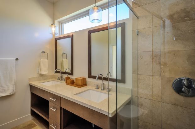 A bright, contemporary bathroom with a long double vanity, two undermount sinks, timber-toned cabinetry, large rectangular mirrors, pendant lighting, and a glass-enclosed shower finished in large beige stone-look tiles.