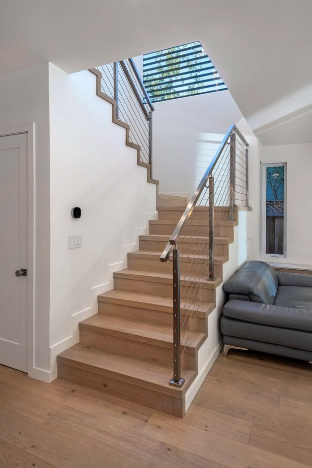 Bright contemporary residential stairway with broad light timber treads and matching timber flooring, white walls, and stainless-steel cable balustrades. A skylight with horizontal exterior louvres brings daylight into the stairwell. Part of a dark grey leather sofa and a narrow window are visible beside the stairs.