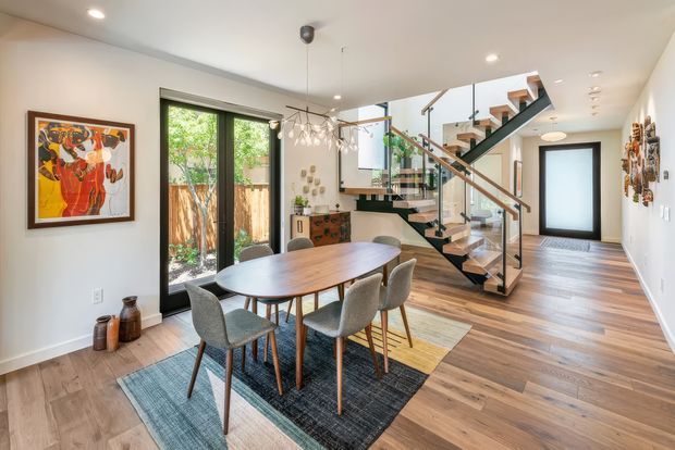 Bright contemporary open-plan dining and entry space with a sculptural timber dining table and upholstered chairs on layered rugs. A glazed, open-riser staircase with timber treads and dark metal framing leads upward beside a timber-floored hallway. Large black-framed glazing opens toward greenery, while recessed ceiling lights and pendant lighting illuminate the neutral interior. Artwork and decorative objects add colour and character.