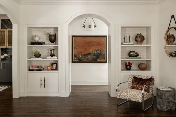 A refined traditional-style interior hallway or entry area with white decorative wall panelling, built-in shelving, arched doorway, dark timber flooring, curated artwork and accessories, and a patterned accent chair beside a small side table. A kitchen or cabinetry area is partially visible through the opening at left.