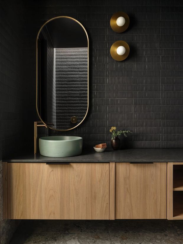A moody, refined bathroom vanity with a large arched bronze-framed mirror, pale green vessel basin, brass wall-mounted tapware, dark tiled walls, dark stone benchtop, and warm timber floating cabinetry. Two round brass-and-white wall lights flank the mirror area, while small plants and accessories add subtle natural detail.