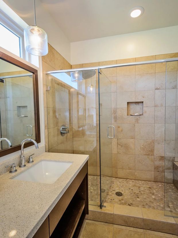 Bright contemporary bathroom featuring a large frameless glass walk-in shower with beige stone-look wall tiles, a pebble-tile shower floor, recessed niche, and chrome fixtures. A wall-mounted mirror, pendant lighting, and a pale engineered-stone vanity benchtop with inset basin and dark timber cabinetry complete the space.