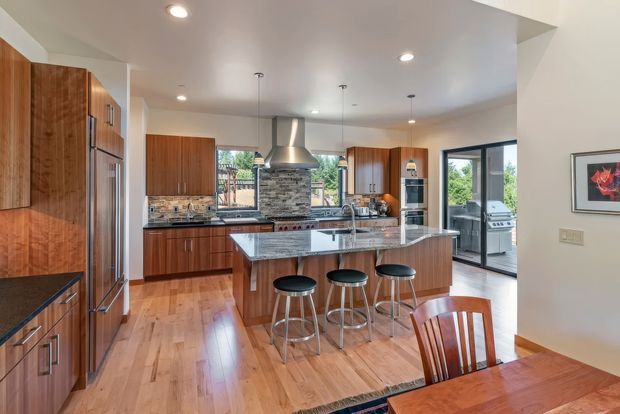 A spacious open-plan kitchen and dining area with warm timber cabinetry, hardwood flooring, stainless-steel appliances, and a large stone island with seating for four. The kitchen features a substantial stone-clad rangehood surround, pendant lighting, recessed ceiling lights, and large glazed doors opening toward an outdoor barbecue area. A dining table occupies the foreground.
