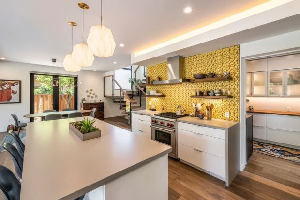 Open-plan kitchen and dining area with a large pale island, white flat-panel cabinetry, timber flooring, and a striking yellow geometric tile splashback. The kitchen includes a stainless-steel rangehood and oven, open timber shelves, pendant lighting, and a walk-in pantry or utility space visible at right. A staircase and glazed doors connect the space to the rest of the home.