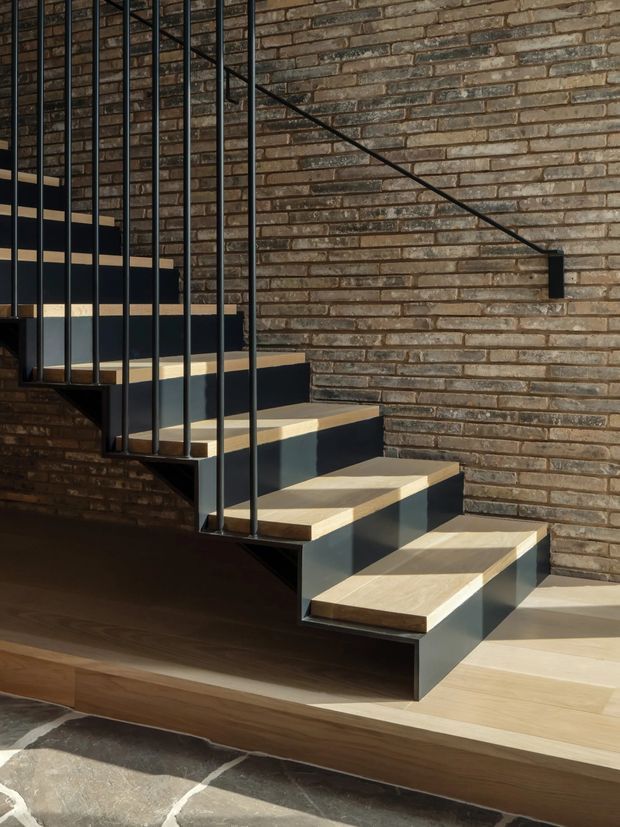 Interior architectural detail of a contemporary floating staircase with pale timber treads, dark painted steel stringers and slender black metal balustrade. The staircase is set against an exposed warm brown brick wall, with a dark timber floor and a large-format stone floor visible at the bottom edge. Strong sunlight creates crisp shadows across the steps and wall.
