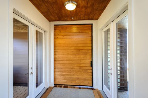 A bright, enclosed entry vestibule with a prominent horizontal timber-clad front door, matching timber ceiling, white-painted walls and trim, and extensive glazed sidelights or doors on both sides. The space has a grey floor mat and warm natural-wood detailing, creating a welcoming transitional entrance.