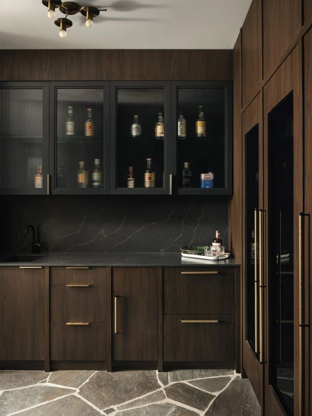 A dark, sophisticated home bar or drinks cabinet with full-height walnut-toned wood joinery, black framed ribbed-glass upper cabinets displaying bottles, a dark veined stone backsplash and benchtop, brass hardware, integrated sink, and tall glass-front storage at right. Irregular natural stone flooring and warm globe lighting add texture and atmosphere.