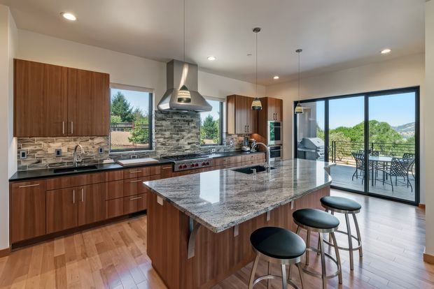 Spacious contemporary kitchen with warm walnut-toned cabinetry, a large polished stone island with seating, stainless-steel appliances, and a stacked-stone rangehood feature wall. Pendant lights and recessed ceiling lighting illuminate the open-plan space, which connects to a glazed outdoor terrace with views beyond.