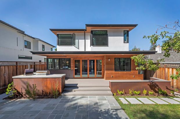 A contemporary two-storey residential rear facade with a warm timber-clad ground floor, white rendered upper level, broad flat roof overhangs, and horizontal louvred window shades. The house opens onto a landscaped patio with large grey paving, raised timber-clad planters or walls, steps, lawn, and a built-in outdoor kitchen or counter. Strong indoor-outdoor connection through extensive dark-framed glazing and timber-framed doors.
